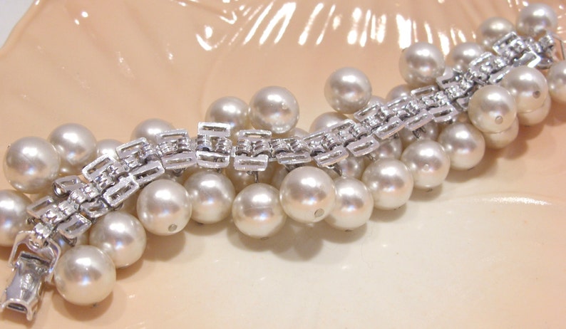 May include: A vintage bracelet featuring a silver-tone metal link design adorned with numerous large, round, creamy white faux pearls. The bracelet is displayed on a light peach-colored surface, showcasing its elegant design.
