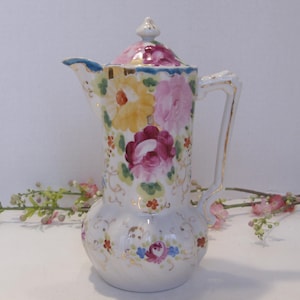 May include: A white porcelain coffee pot with a lid, decorated with colorful floral patterns. The flowers are in shades of pink, yellow, and burgundy, with gold accents on the handle and rim. The pot has a bulbous base and a slender spout.