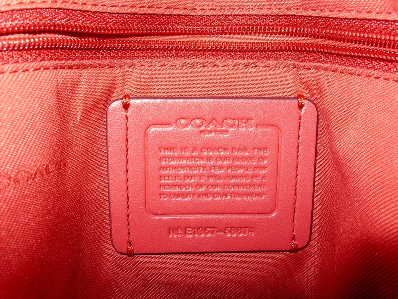 May include: Close-up of the interior of a red Coach bag. The image shows a red leather tag with the Coach logo and text, stitched onto the red fabric lining. A red zipper is visible at the top of the image.
