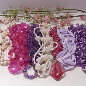 May include: A collection of six necklaces featuring a variety of materials including white seashells, translucent pink cylindrical beads, deep purple faceted beads, white textured spherical beads, light lavender rectangular beads, and purple beaded clusters.