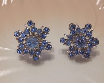 May include: A pair of vintage snowflake-shaped earrings. Each earring features a central light blue gemstone surrounded by smaller, matching blue stones, creating a sparkling, floral design. The earrings are set in a silver-toned metal.