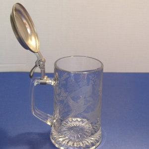 May include: Clear glass beer stein with a hinged metal lid. The stein features an etched design of birds and foliage. The handle is rectangular, and the base has a decorative, ridged pattern.