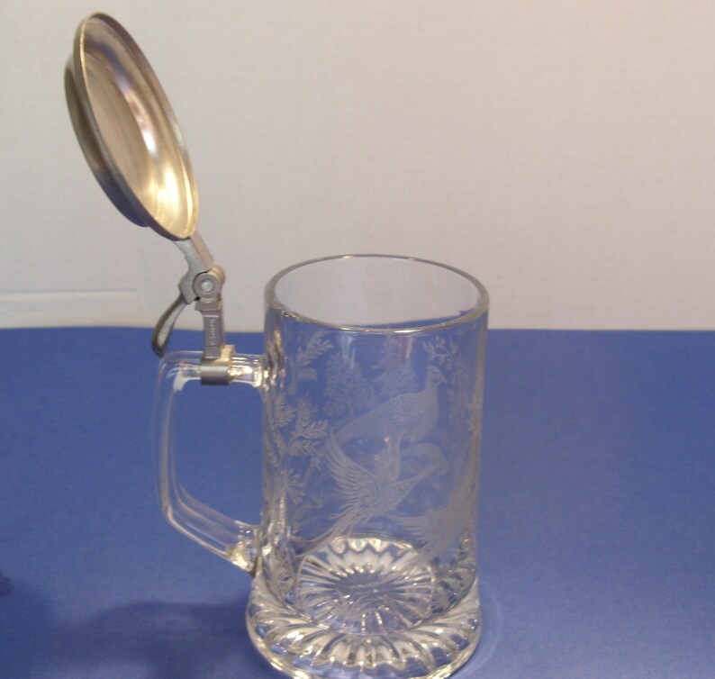 May include: Clear glass beer stein with a hinged metal lid. The stein features an etched design of birds and foliage. The handle is rectangular, and the base has a decorative, ridged pattern.