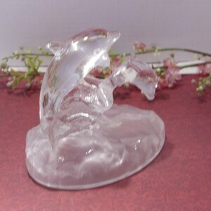 May include: A clear glass dolphin sculpture. Two dolphins are depicted leaping from the water. The sculpture is set on a rounded base. The background features pink and green floral accents on a maroon surface.