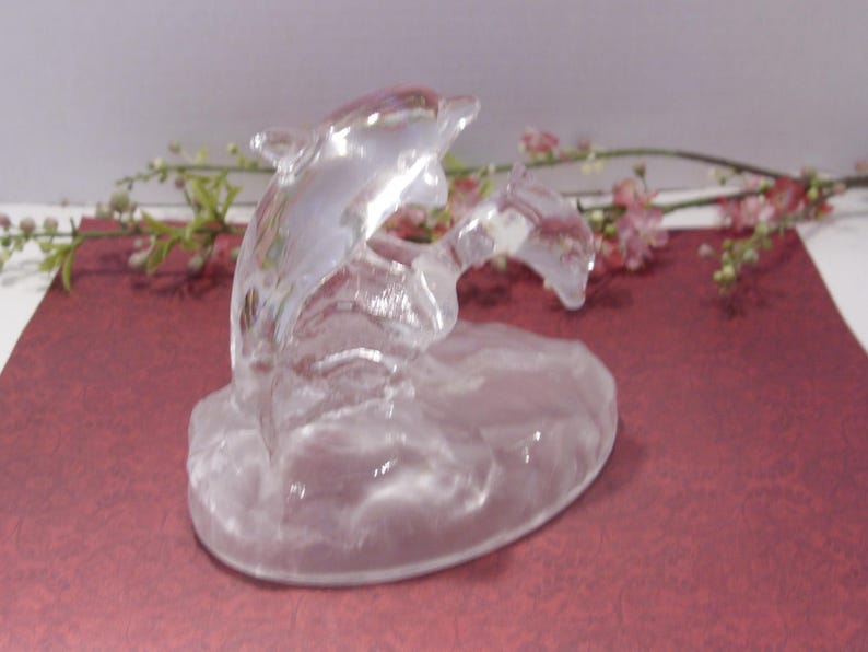 May include: A clear glass dolphin sculpture. Two dolphins are depicted leaping from the water. The sculpture is set on a rounded base. The background features pink and green floral accents on a maroon surface.