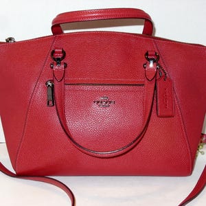 May include: A red leather handbag with a top handle and a detachable shoulder strap. The bag features a front zippered pocket, a small tag, and silver-tone hardware. The Coach logo is visible on the front of the bag.