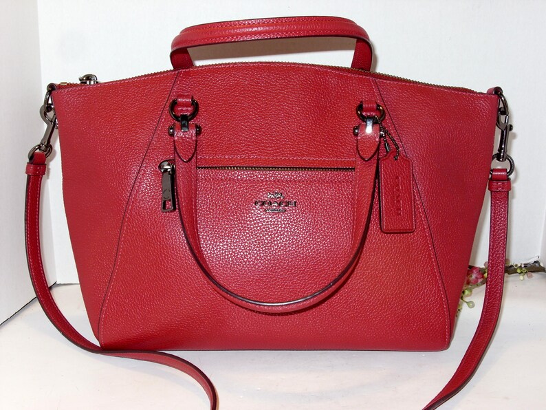 May include: A red leather handbag with a top handle and a detachable shoulder strap. The bag features a front zippered pocket, a small tag, and silver-tone hardware. The Coach logo is visible on the front of the bag.