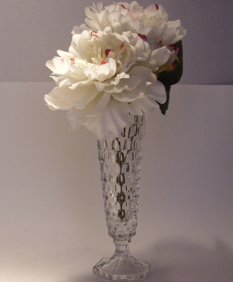 Puede incluir: Un jarr&oacute;n de vidrio transparente y texturizado contiene un ramo de peon&iacute;as blancas con toques de rojo. El jarr&oacute;n tiene una base hexagonal y un cuerpo alto y estampado. Las flores son llenas y exuberantes, creando un arreglo floral cl&aacute;sico.