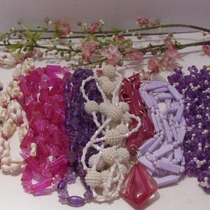 May include: A collection of six necklaces featuring various designs including white shell beads, translucent pink cylindrical beads, deep purple faceted beads, white textured spherical beads, light lavender rectangular beads, and purple floral beads.