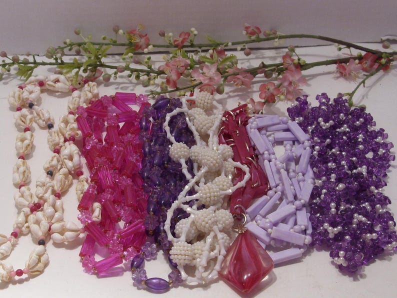 May include: A collection of six necklaces featuring various designs including white shell beads, translucent pink cylindrical beads, deep purple faceted beads, white textured spherical beads, light lavender rectangular beads, and purple floral beads.