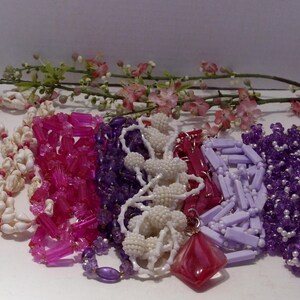 May include: A collection of six necklaces featuring various designs, including white seashell strands, translucent hot pink cylindrical beads, deep purple faceted beads, white textured spherical beads, a red diamond-shaped pendant, and light purple rectangular beads.