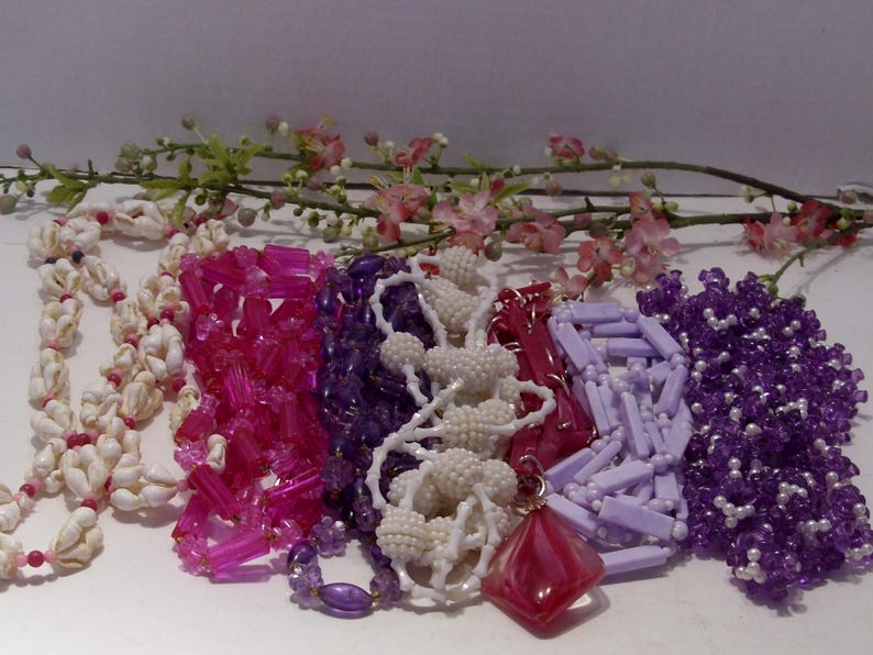 May include: A collection of six necklaces featuring various designs, including white seashell strands, translucent hot pink cylindrical beads, deep purple faceted beads, white textured spherical beads, a red diamond-shaped pendant, and light purple rectangular beads.