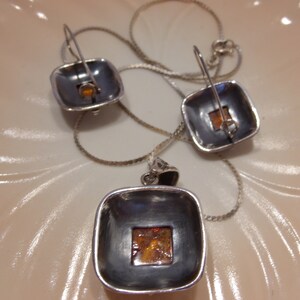 K&ouml;nnte beinhalten: Ein silbernes Halsketten- und Ohrring-Set mit quadratischen Bernstein-Steinen. Die Halskette hat eine silberne Kette und einen quadratischen Anh&auml;nger mit einem Bernstein-Stein in der Mitte. Die Ohrringe sind silberne Creolen mit quadratischen Bernstein-Steinen, die an der Unterseite befestigt sind.