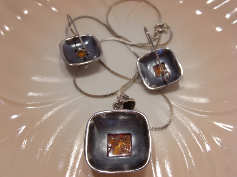 K&ouml;nnte beinhalten: Ein silbernes Halsketten- und Ohrring-Set mit quadratischen Bernstein-Steinen. Die Halskette hat eine silberne Kette und einen quadratischen Anh&auml;nger mit einem Bernstein-Stein in der Mitte. Die Ohrringe sind silberne Creolen mit quadratischen Bernstein-Steinen, die an der Unterseite befestigt sind.