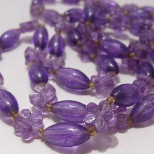 May include: A necklace featuring a repeating pattern of translucent purple oval beads and light purple flower-shaped beads. The beads are strung together with small, clear spacers, creating a continuous strand of violet-toned floral and oval elements.