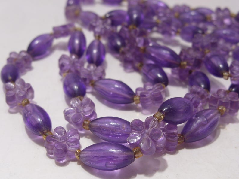 May include: A necklace featuring a repeating pattern of translucent purple oval beads and light purple flower-shaped beads. The beads are strung together with small, clear spacers, creating a continuous strand of violet-toned floral and oval elements.