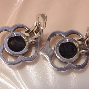 May include: A pair of silver tone clip-on earrings with a floral design and black stones in the center.