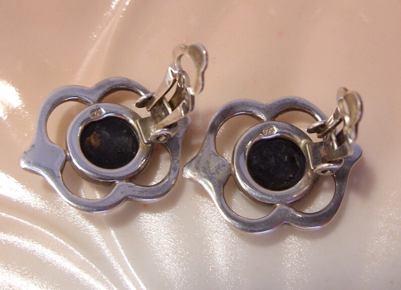 May include: A pair of silver tone clip-on earrings with a floral design and black stones in the center.