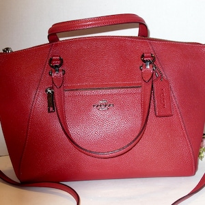 May include: A red leather handbag with a top handle and a detachable shoulder strap. The bag features a front pocket with a zipper and a small tag. The hardware is silver-toned.