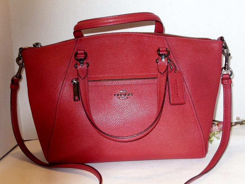May include: A red leather handbag with a top handle and a detachable shoulder strap. The bag features a front pocket with a zipper and a small tag. The hardware is silver-toned.