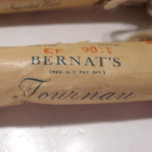 May include: Close-up of a vintage Bernat's Tournai yarn label. The label is tan with blue and orange floral accents. The text "BERNAT'S" and "Tournai" are visible. The label also includes the text "(REG. U.S. PAT. OFF.)".