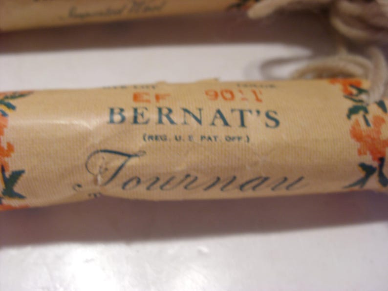 May include: Close-up of a vintage Bernat's Tournai yarn label. The label is tan with blue and orange floral accents. The text "BERNAT'S" and "Tournai" are visible. The label also includes the text "(REG. U.S. PAT. OFF.)".