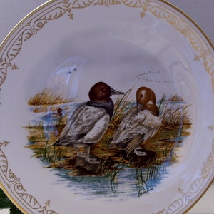 May include: A white ceramic plate with a gold rim and a detailed design. The plate features a painting of two ducks in a marsh setting. The ducks are brown and white with a detailed design. The plate is signed "Boehm" in the bottom right corner.