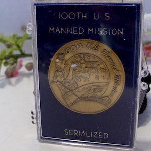 May include: A navy blue commemorative plaque with gold lettering and a gold-colored medallion. The text reads "100th U.S. Manned Mission" and "Serialized". The medallion features an illustration of a space scene.