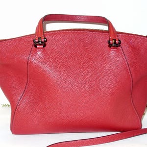 May include: A red leather handbag with a top handle and a detachable shoulder strap. The bag has a textured surface, a zipper closure, and dark metal hardware. The bag's design is a classic, structured style.
