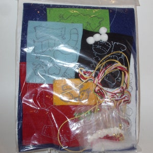 May include: A craft kit containing felt pieces in various colors, including blue, red, green, and black. The kit includes white pom-poms, embroidery floss, and a small piece of felt with text. The felt pieces have printed patterns for cutting and sewing.