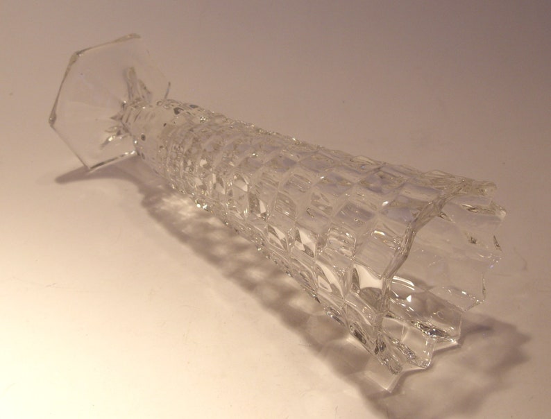 Puede incluir: Un jarr&oacute;n de vidrio transparente con un patr&oacute;n geom&eacute;trico texturizado. El jarr&oacute;n tiene una base hexagonal ensanchada y un cuerpo largo y cil&iacute;ndrico. El vidrio es transparente y est&aacute; ligeramente inclinado hacia la izquierda.