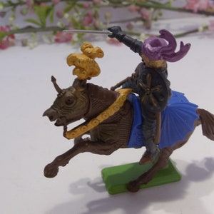 May include: A plastic toy knight riding a brown horse with a gold and blue saddle. The knight is wearing a purple helmet with a gold cross on his chest and is holding a sword.
