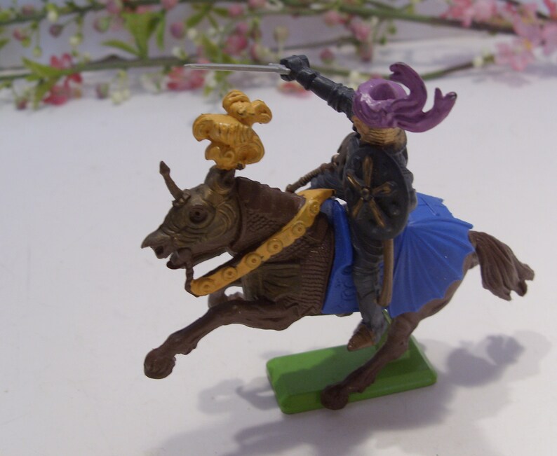May include: A plastic toy knight riding a brown horse with a gold and blue saddle. The knight is wearing a purple helmet with a gold cross on his chest and is holding a sword.