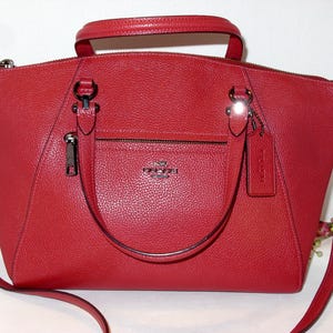 May include: Red leather handbag with a top handle and a detachable shoulder strap. The bag has a front zipper pocket and a small tag. The hardware is silver-toned. The bag has a textured finish.