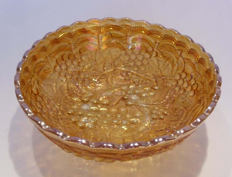 Puede incluir: Un cuenco de vidrio &aacute;mbar iridiscente con borde festoneado. El cuenco presenta un dise&ntilde;o en relieve de uvas y hojas. El vidrio tiene una cualidad brillante y opalescente, reflejando la luz con una gama de colores. Objeto decorativo.