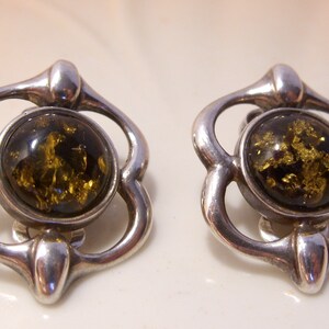 May include: A pair of silver earrings with a green amber stone in the center of each earring. The earrings are in the shape of a flower with four petals.
