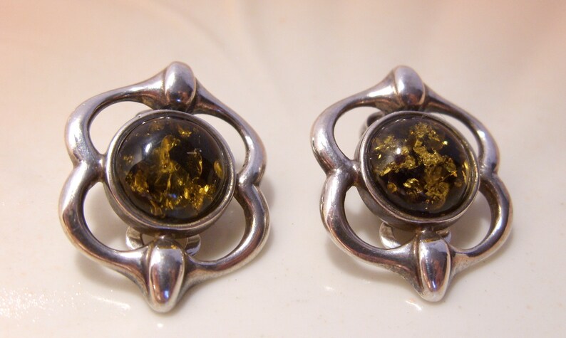 May include: A pair of silver earrings with a green amber stone in the center of each earring. The earrings are in the shape of a flower with four petals.