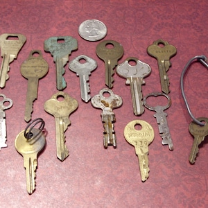 May include: A collection of antique keys in various shapes and sizes, made of brass and silver-toned metals. Some keys show signs of age and wear, with a quarter coin placed among them. The keys are arranged on a textured, maroon surface.