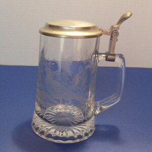 May include: Clear glass beer stein with a hinged metal lid and handle. The glass is etched with a pheasant and foliage design. The base of the stein has a decorative, scalloped edge.
