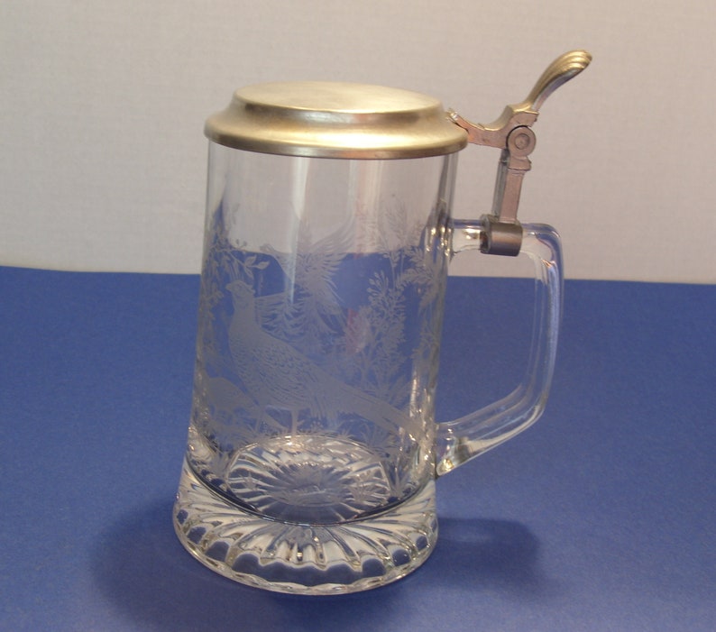May include: Clear glass beer stein with a hinged metal lid and handle. The glass is etched with a pheasant and foliage design. The base of the stein has a decorative, scalloped edge.