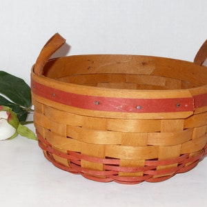 May include: A woven basket with two handles, made of natural wood with a red painted trim. The basket is empty and sitting on a white surface.