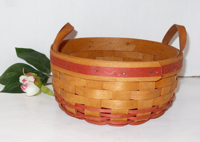 May include: A woven basket with two handles, made of natural wood with a red painted trim. The basket is empty and sitting on a white surface.
