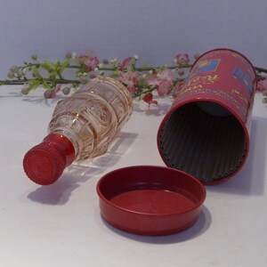 May include: A clear glass bottle with a red screw cap, positioned next to a cylindrical red metal container and its matching red lid. The bottle features a textured, embossed surface pattern, while the container has visible text and graphics.