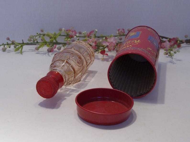 May include: A clear glass bottle with a red screw cap, positioned next to a cylindrical red metal container and its matching red lid. The bottle features a textured, embossed surface pattern, while the container has visible text and graphics.