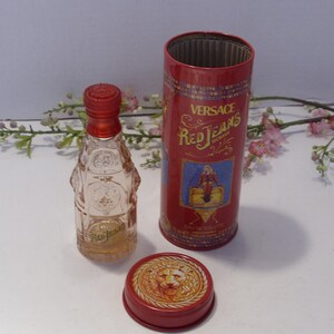 May include: A clear glass perfume bottle with a red cap, a matching red cylindrical metal canister, and a separate red lid featuring a raised lion head emblem. The canister displays the text Versace Red Jeans and an illustration of a figure.