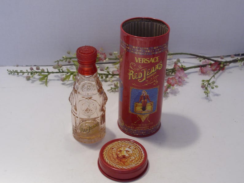 May include: A clear glass perfume bottle with a red cap, a matching red cylindrical metal canister, and a separate red lid featuring a raised lion head emblem. The canister displays the text Versace Red Jeans and an illustration of a figure.