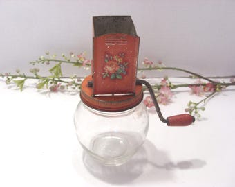 May include: Vintage red and orange hand-crank food grinder with a floral design on the front. The grinder sits atop a clear glass jar with a metal lid. The handle is made of wood and metal.