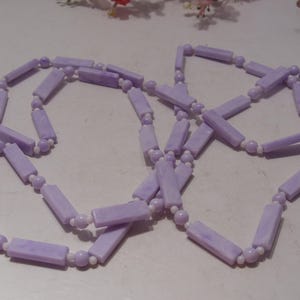 May include: A long necklace featuring alternating light purple rectangular beads and small white spherical beads. The rectangular beads have a marbled texture, and the necklace is arranged in a loose, overlapping loop pattern.