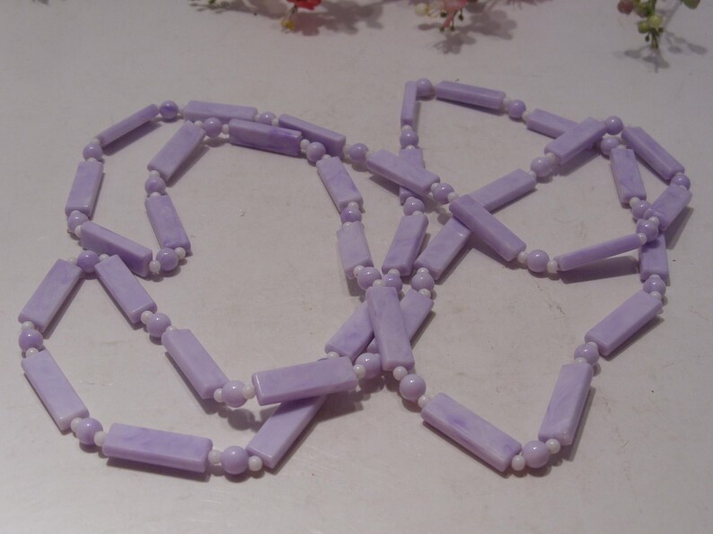 May include: A long necklace featuring alternating light purple rectangular beads and small white spherical beads. The rectangular beads have a marbled texture, and the necklace is arranged in a loose, overlapping loop pattern.