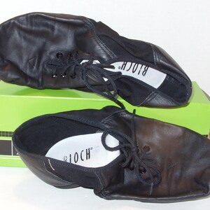 Puede incluir: Un par de zapatos de baile de cuero negro con cordones, apoyados en una caja verde. Los zapatos tienen una puntera redondeada y una suela blanda, con la marca "BLOCH" visible en la plantilla. Zapatos de baile.